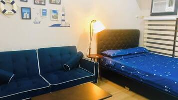 A room with a blue sofa, a small table, a bed with a blue bedspread, a wall-mounted air conditioner, and various framed pictures on the wall.