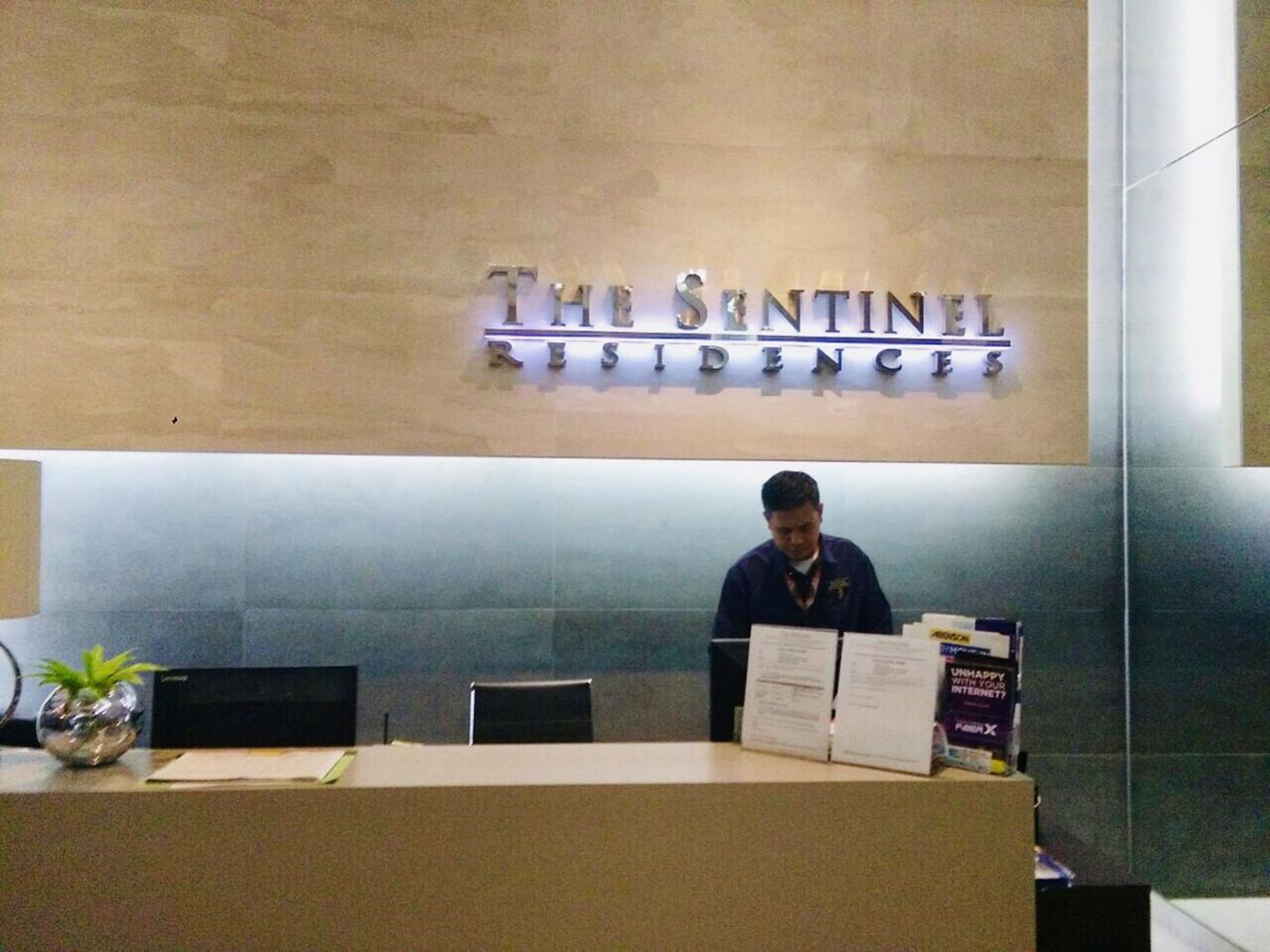 A reception desk at The Sintonel Residences with a man standing behind it, a sign above the desk, and some documents on the counter.