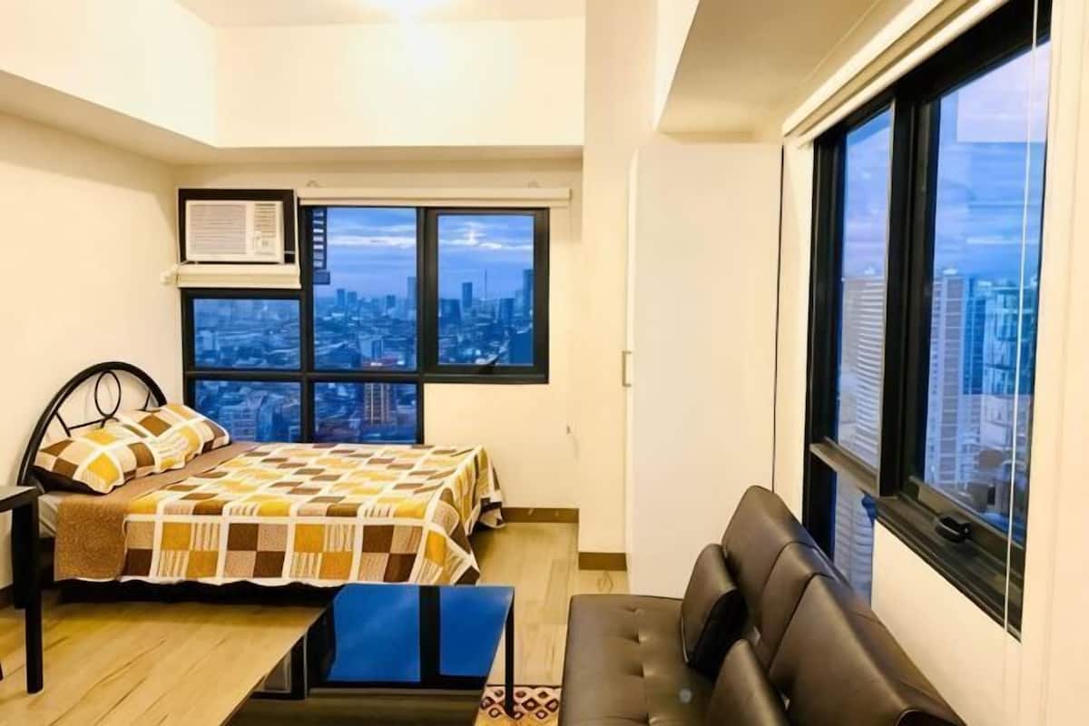A bedroom with a bed, a desk, a chair, a window with a city view, and an air conditioning unit.