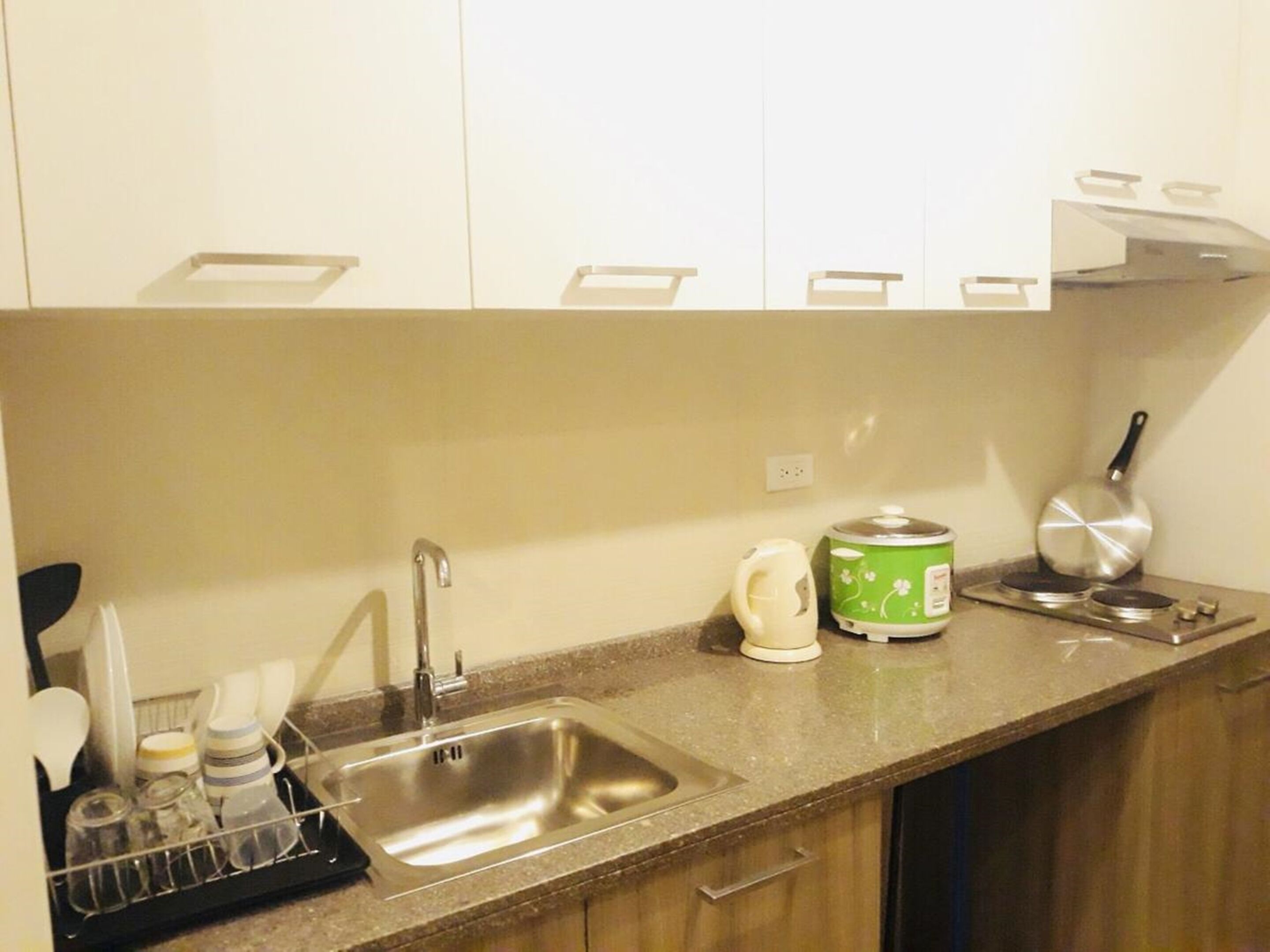 A kitchen with a stainless steel sink, a kettle, a frying pan, and a green electric kettle on the countertop.