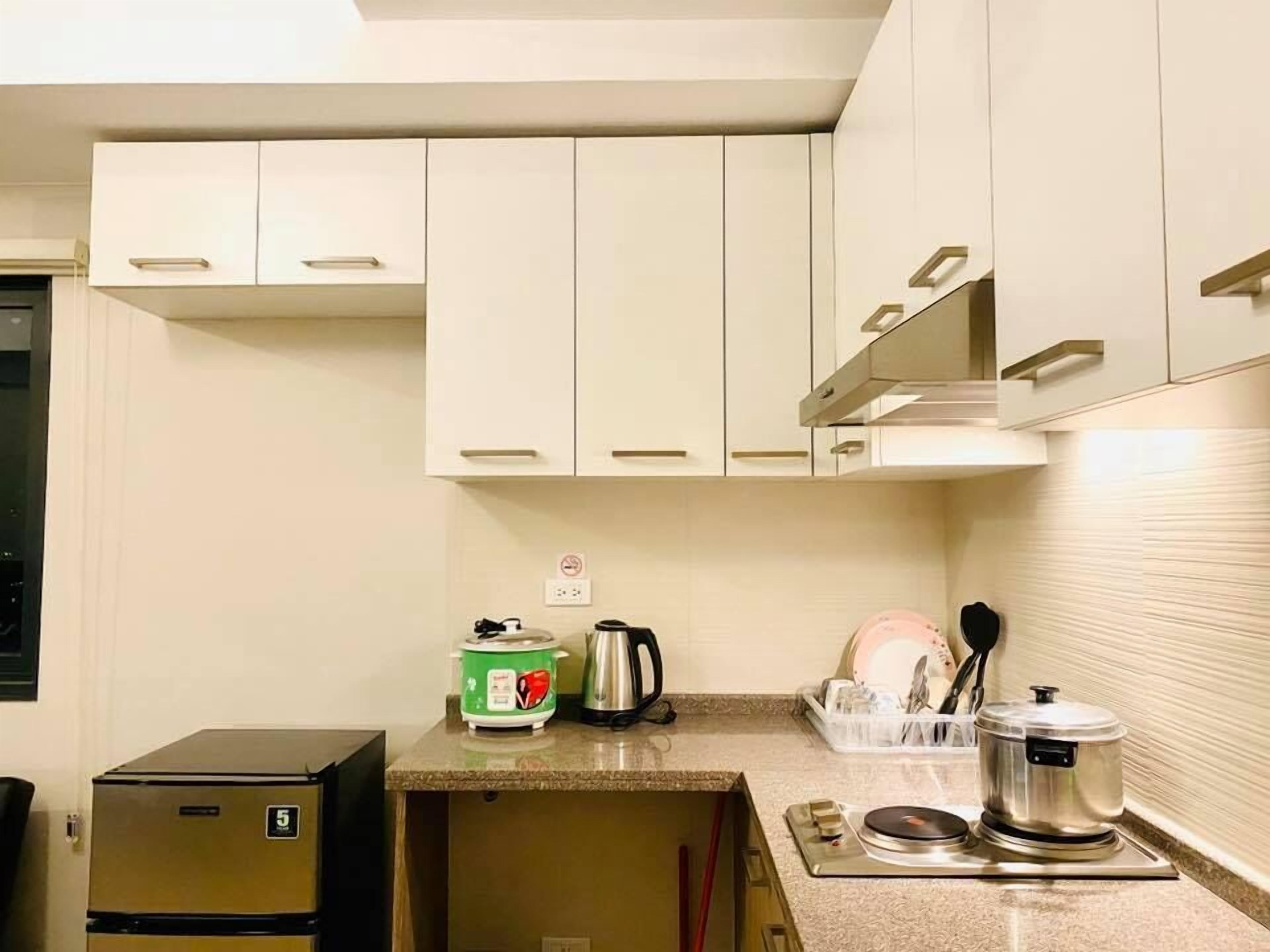 Una cocina compacta con armarios blancos, un refrigerador pequeño, una tetera, un escurreplatos y una estufa portátil.