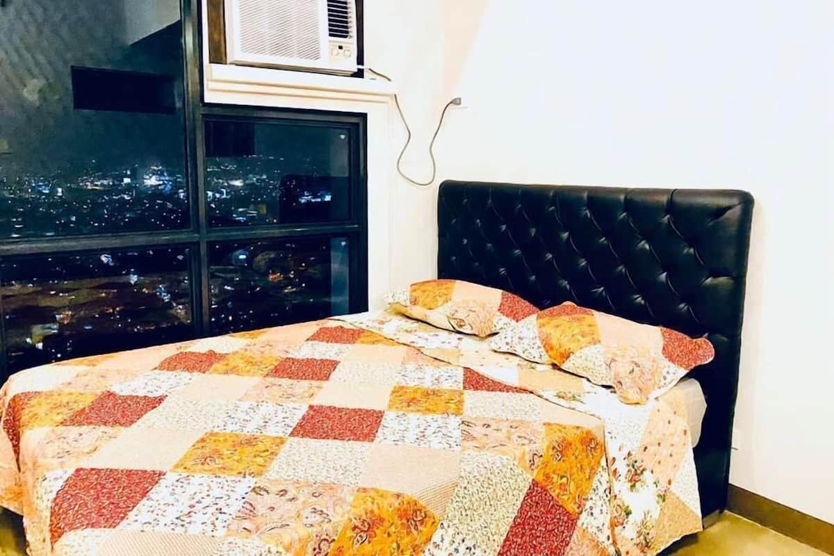 A bedroom with a checkered bedspread, a black upholstered headboard, and a window with a city view.