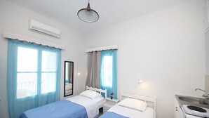 A hotel room with two single beds, a small bedside table with a lamp, a mirror, a window with curtains, an air conditioning unit, and a kitchenette with a sink and a microwave. - Twin Shades (Kalymnos)
