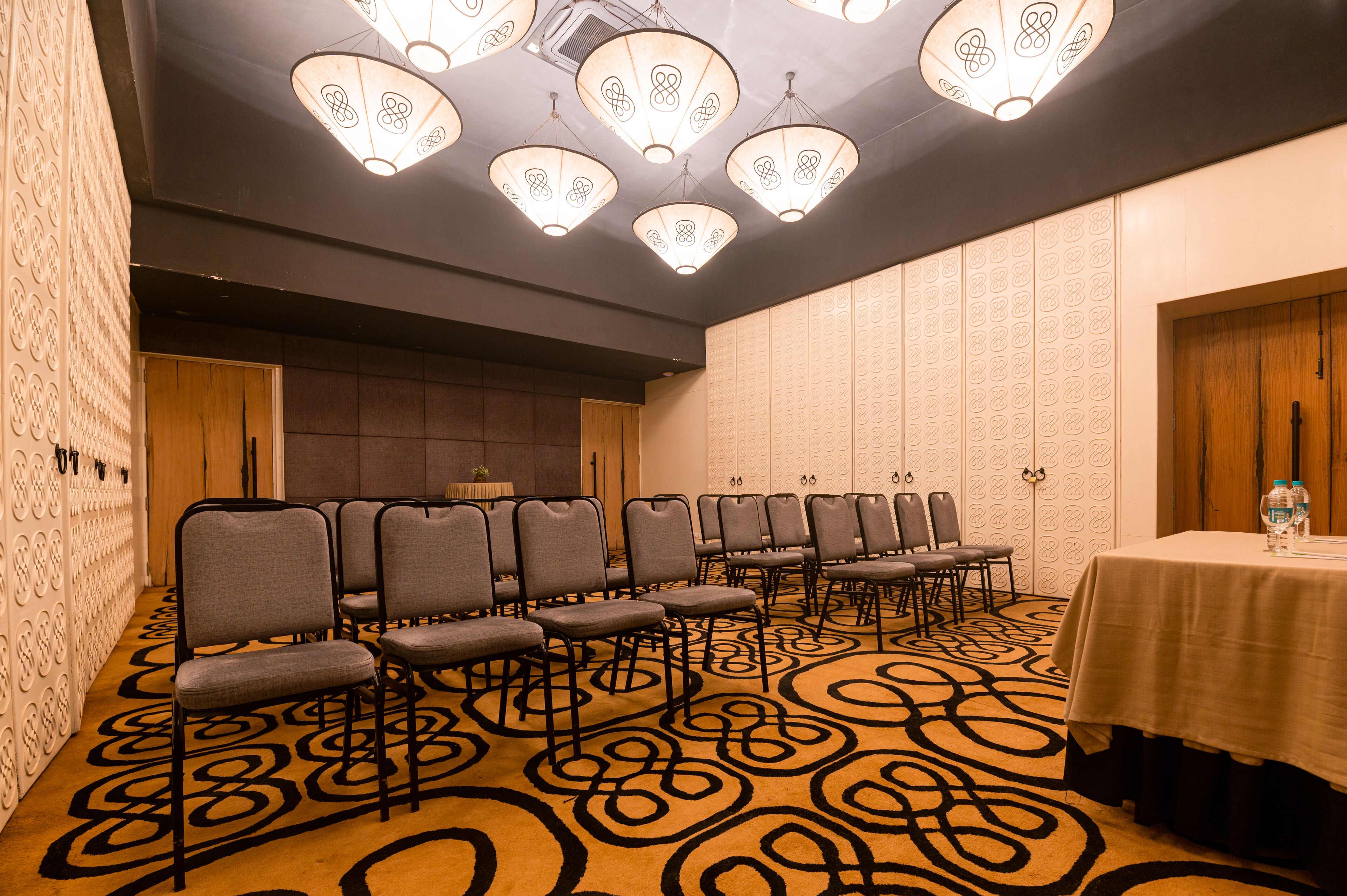 Un salón de conferencias con filas de sillas, una mesa con botellas de agua, alfombra estampada y luces decorativas en el techo.