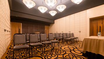 Un salón de conferencias con filas de sillas, una mesa con botellas de agua, alfombra estampada y luces decorativas en el techo.