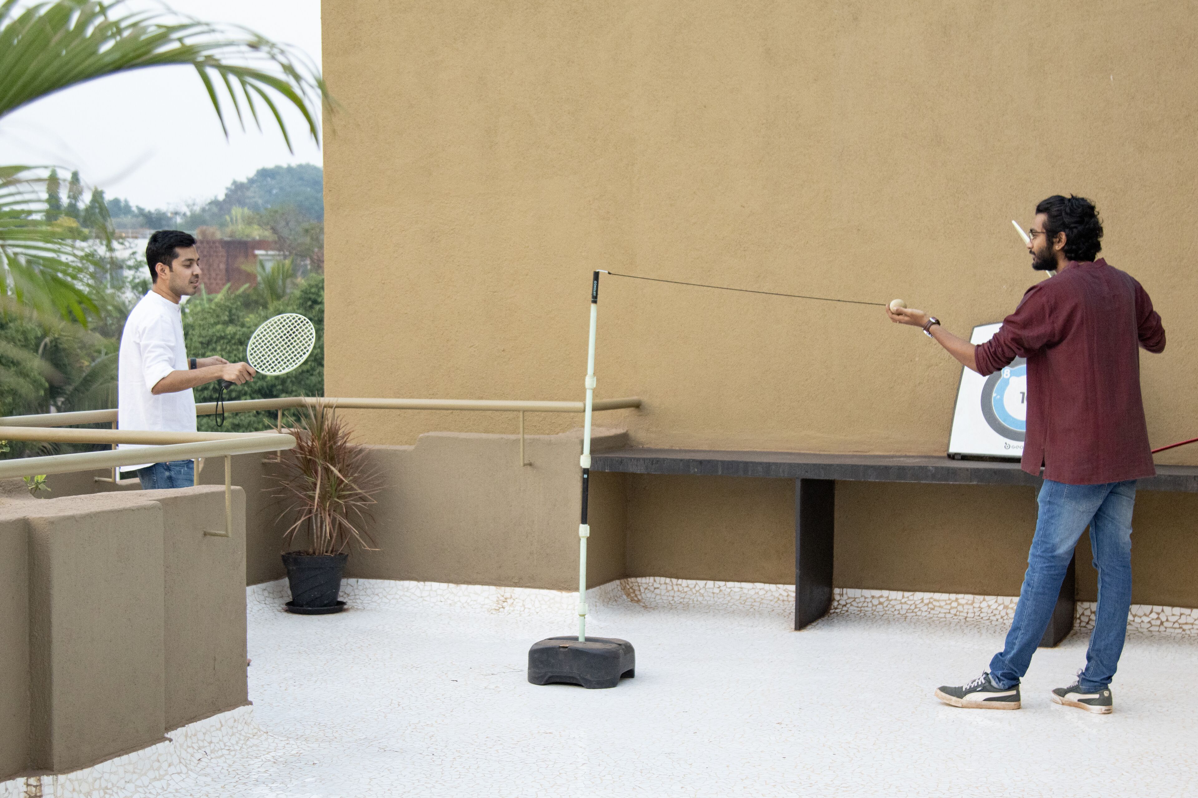 Un hombre juega al bádminton en una terraza en la azotea, con una maceta y una mesa al fondo.