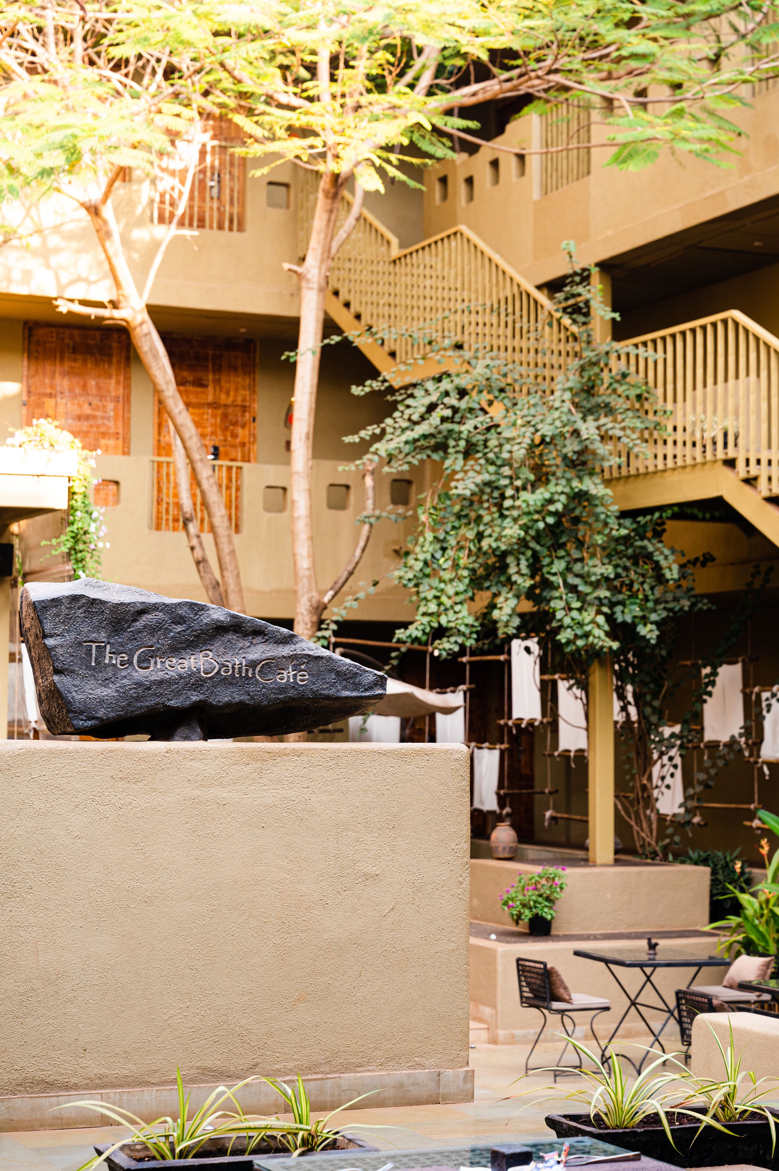 A courtyard with a stone bench labeled "The Great Bath Cafe," surrounded by potted plants and outdoor seating.