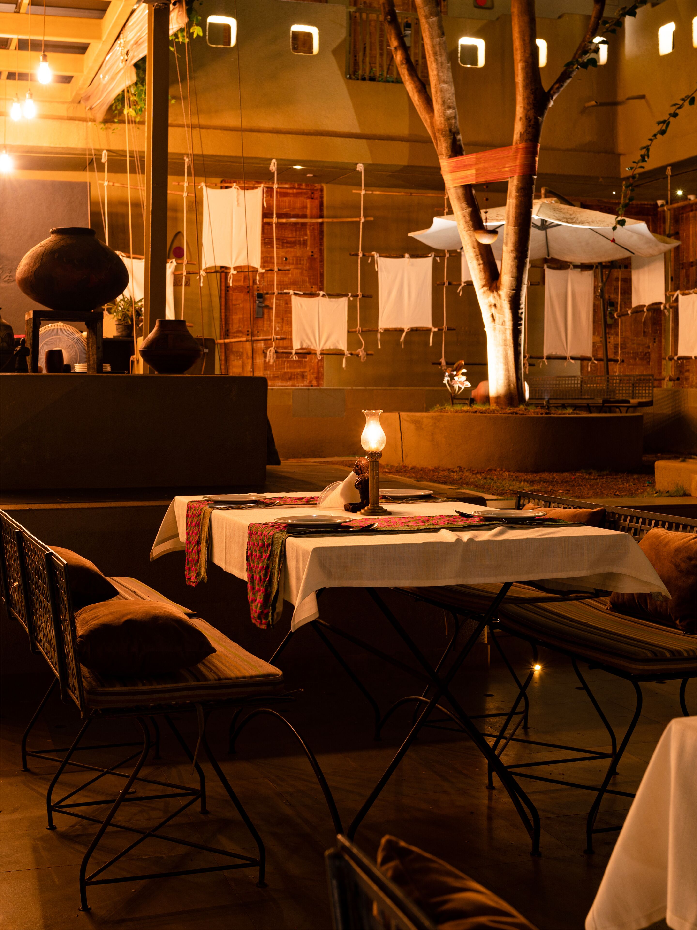 An outdoor dining area with tables set for two, surrounded by trees and a brick wall.