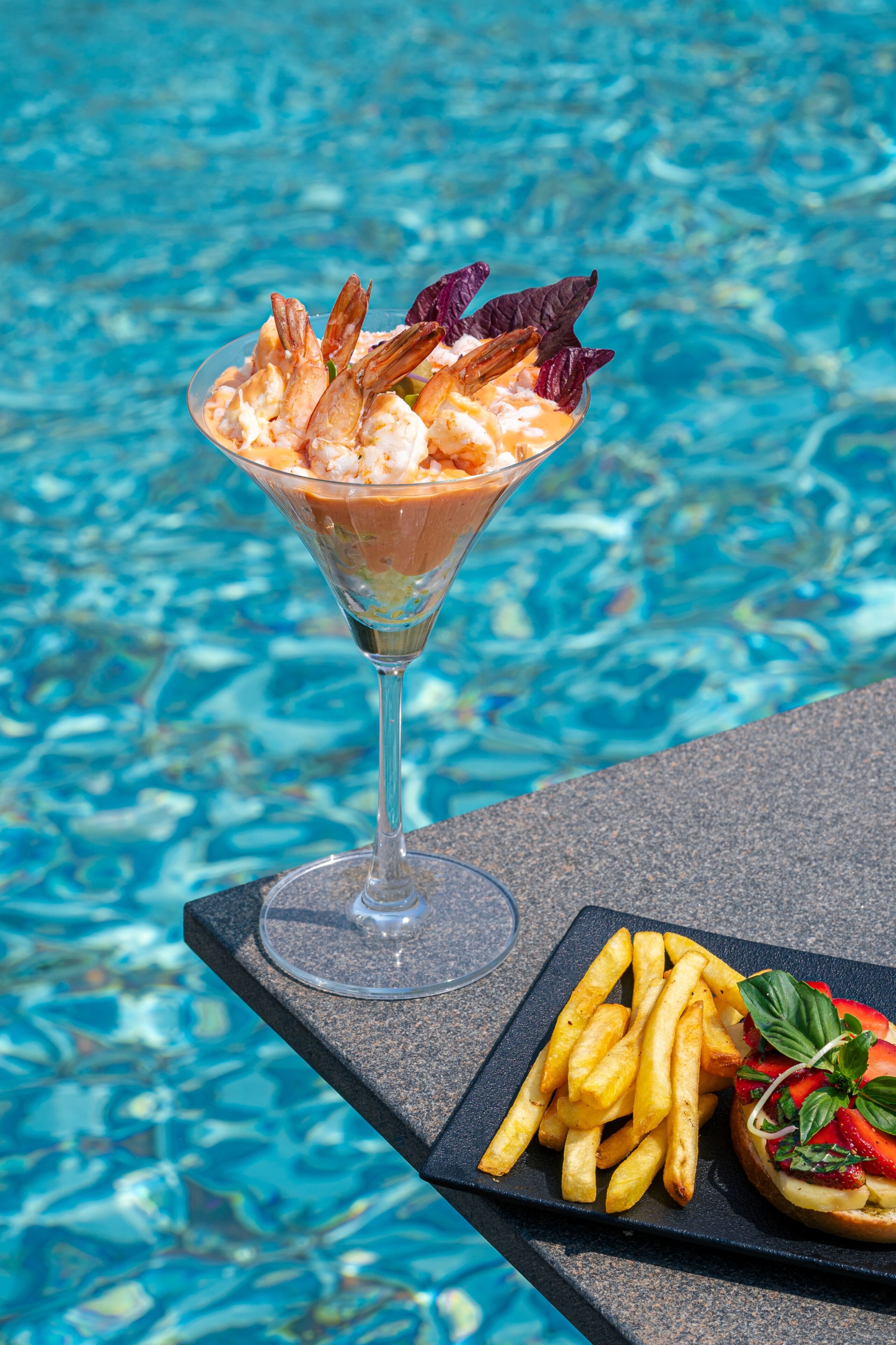 A martini glass with shrimp and a salad on a table by a pool.