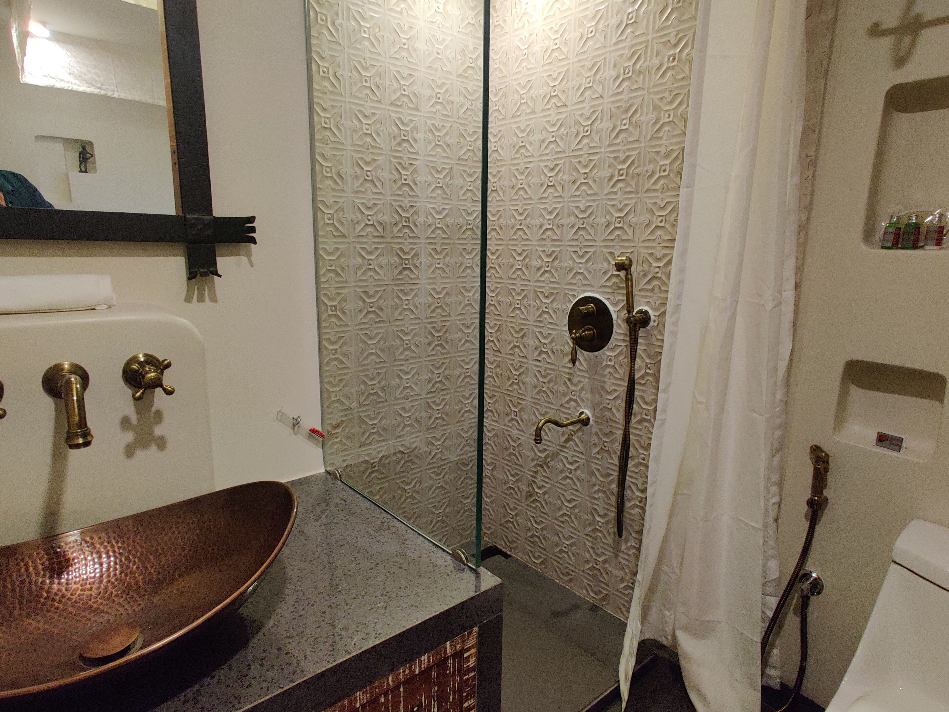 A bathroom with a brown ceramic sink, a shower with a patterned wall, and a toilet with a shelf above it.