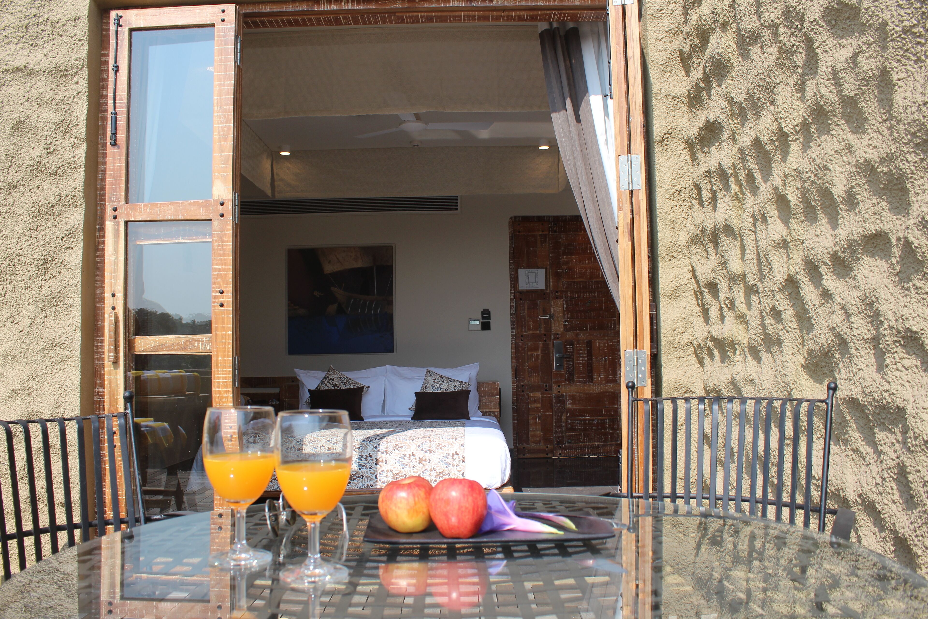 A glass table with two orange drinks, a plate with two apples, and a bed with pillows in the background.