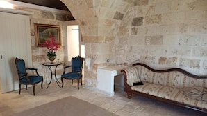 Una habitación con paredes de piedra, un sofá de madera, dos sillones azules y una mesita con un jarrón de flores.