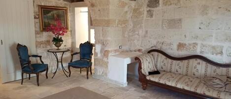 A room with stone walls, a wooden sofa, two blue armchairs, and a small table with a vase of flowers.