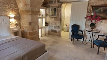 Ein Zimmer mit Steinwänden, ausgestattet mit einem Bett, einem Sessel, einem kleinen Tisch und einer Tür zu einem anderen Raum.