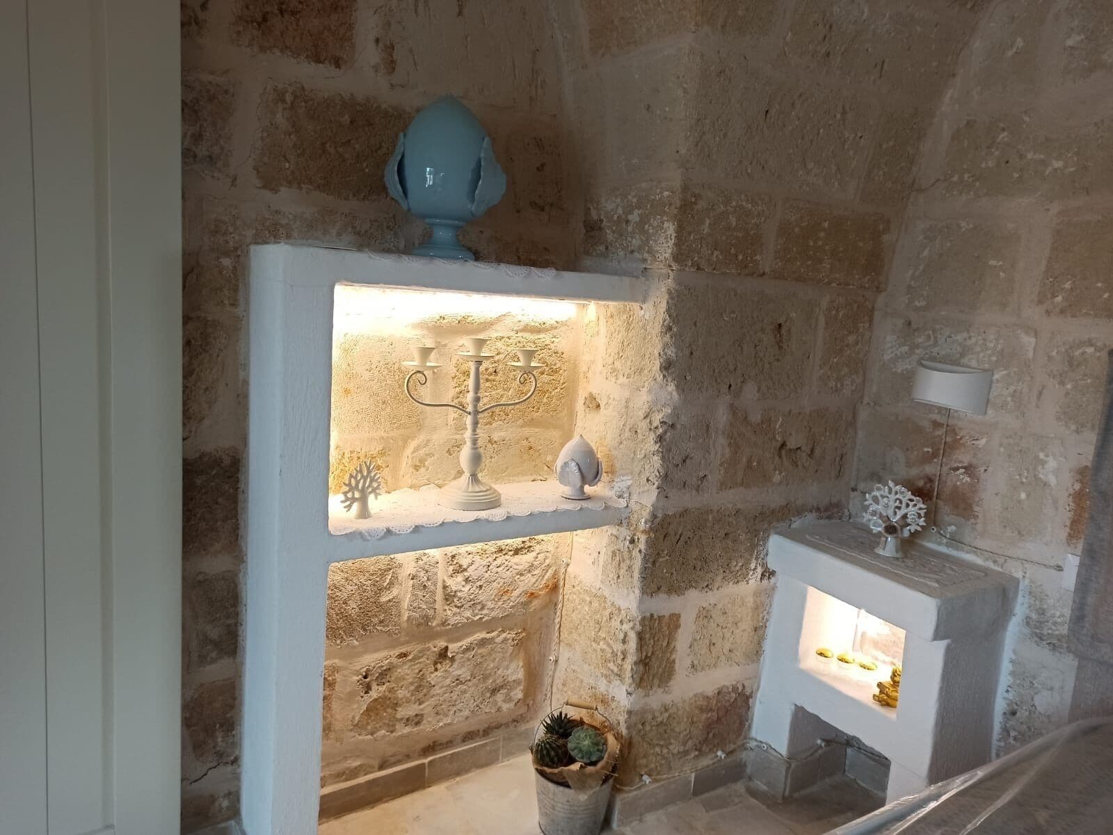 Una pared de piedra rústica con un estante blanco que contiene un candelabro, un pequeño árbol y un objeto decorativo.