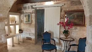 Una habitación con paredes de piedra, una puerta blanca, dos sillones tapizados en azul, una pequeña mesa redonda con una planta en maceta y un cuadro en la pared.