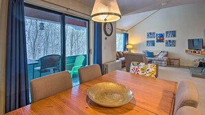 A dining area with a wooden table, chairs, and a decorative bowl. There is a dining table with a bowl in the center, surrounded by chairs. The room has a large window with curtains, a clock on the wall, and a sofa with cushions. - Loon Mountain Condo w/ Pool & Game Room Access! (Lincoln)