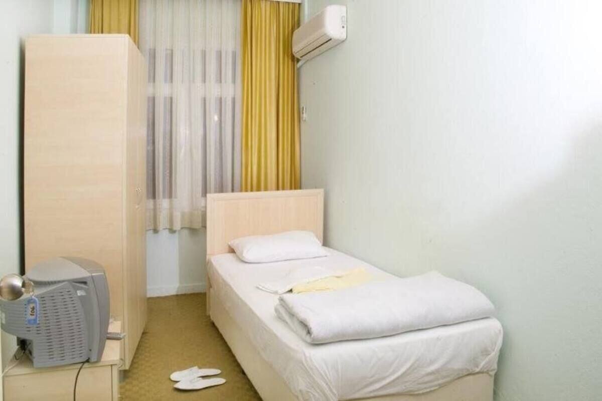 Pequeña habitación individual con una cama y cabecera de madera, una mesita de noche con lámpara, una ventana con cortinas y un equipo de aire acondicionado en la pared.