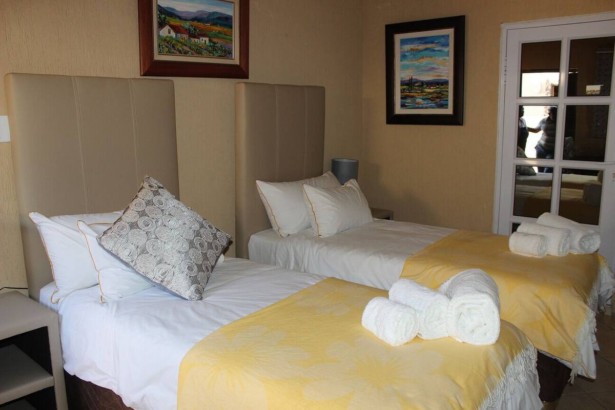 Une chambre d’hôtel avec deux lits, chacun recouvert de draps blancs et d’une couvre-lit à motifs floraux jaunes. Des serviettes sont posées sur les lits, et la chambre dispose d’une fenêtre donnant sur l’extérieur.