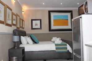 Une chambre d’hôtel comprenant un lit, une table de chevet, une lampe, un tableau encadré, un réfrigérateur et un téléviseur fixé au mur.
