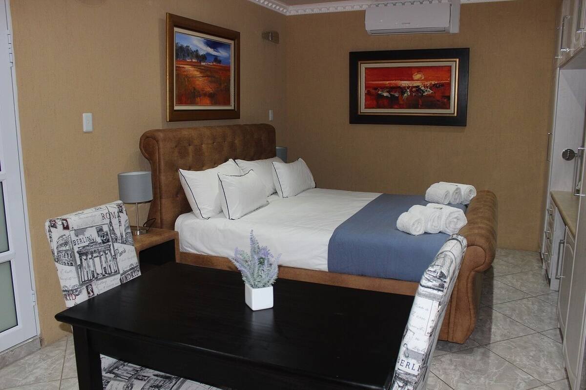 Une chambre d’hôtel avec un lit, une table de chevet ornée d’une plante, une chaise avec un coussin représentant un paysage urbain, et deux tableaux encadrés accrochés au mur.