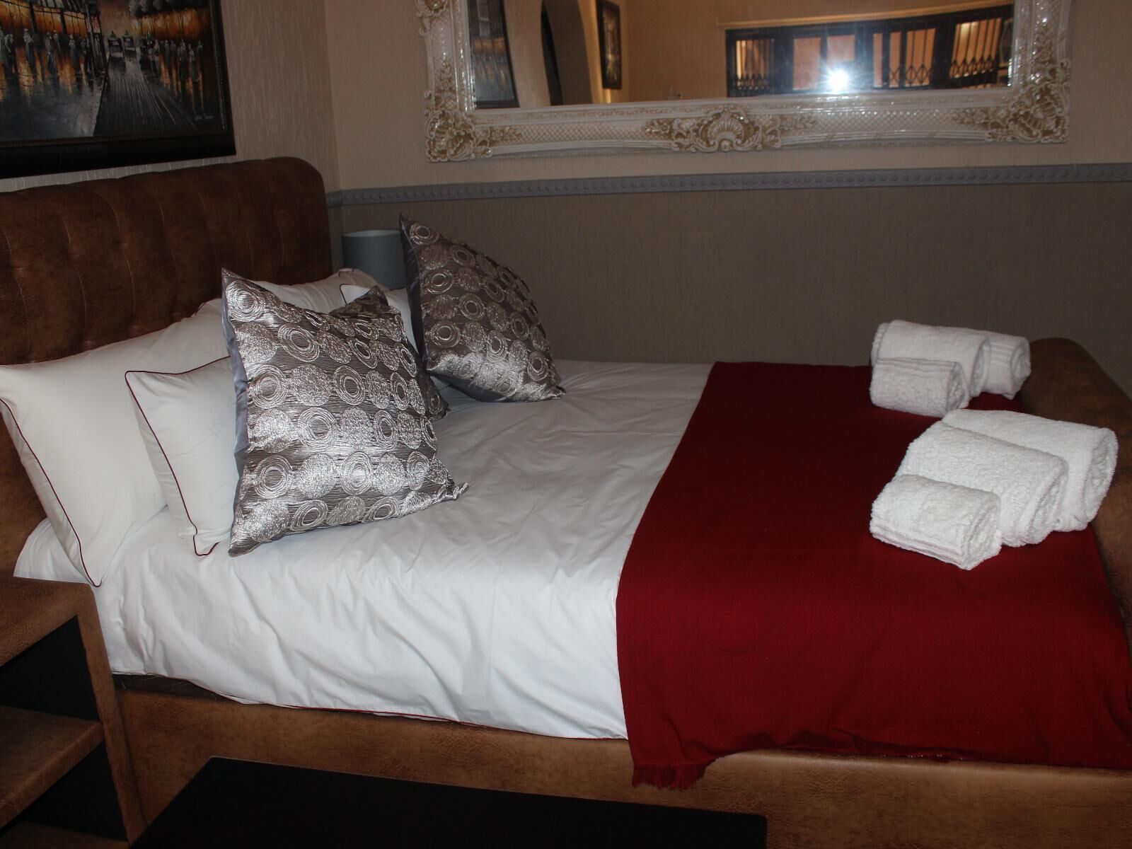 A neatly made bed with white linens, two silver pillows, a red throw blanket, and white towels on a wooden headboard.