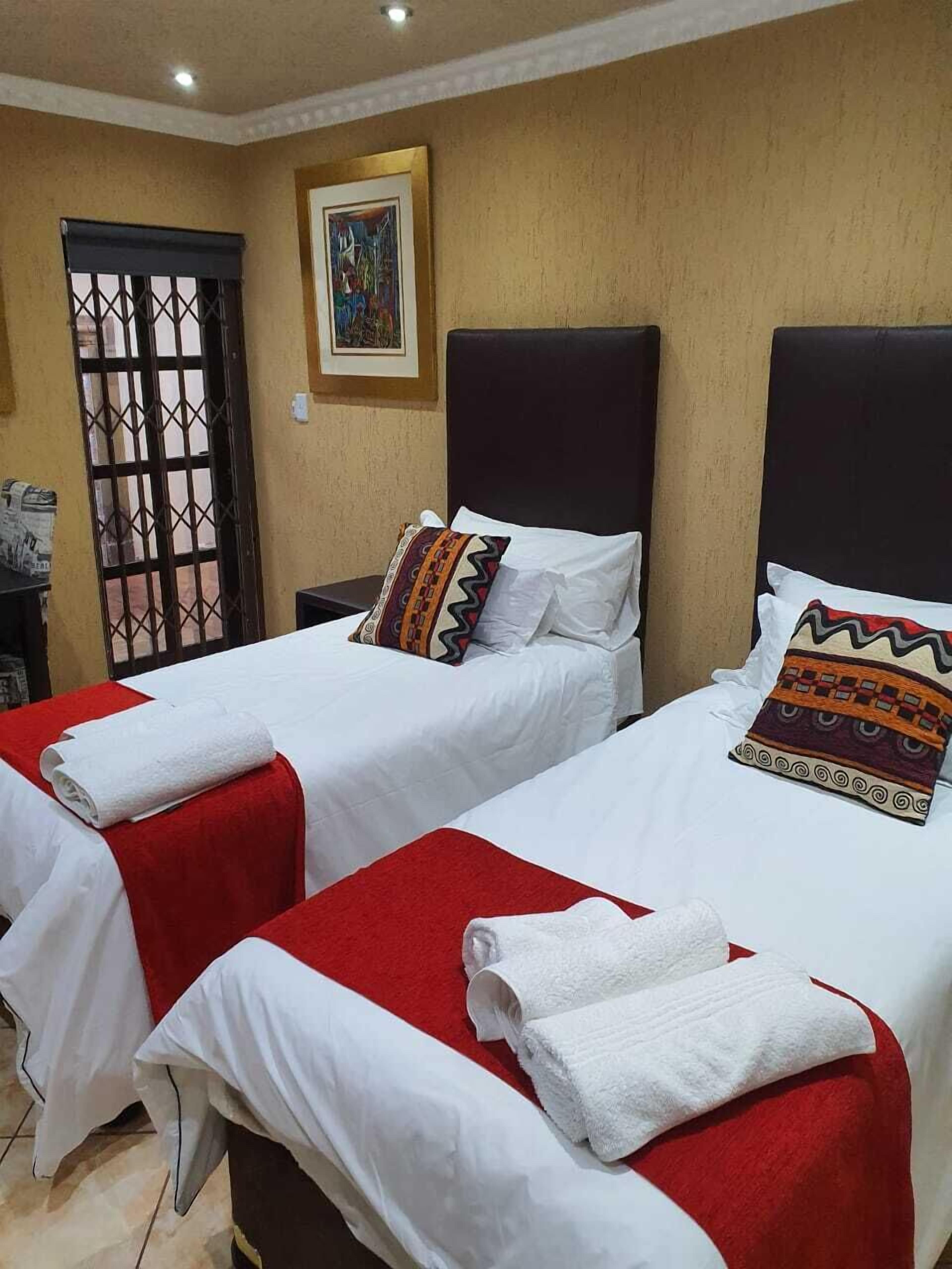 A hotel room with two beds, each with a red bedspread and white linens. There is a painting on the wall above the beds and a door with a metal gate to the left.