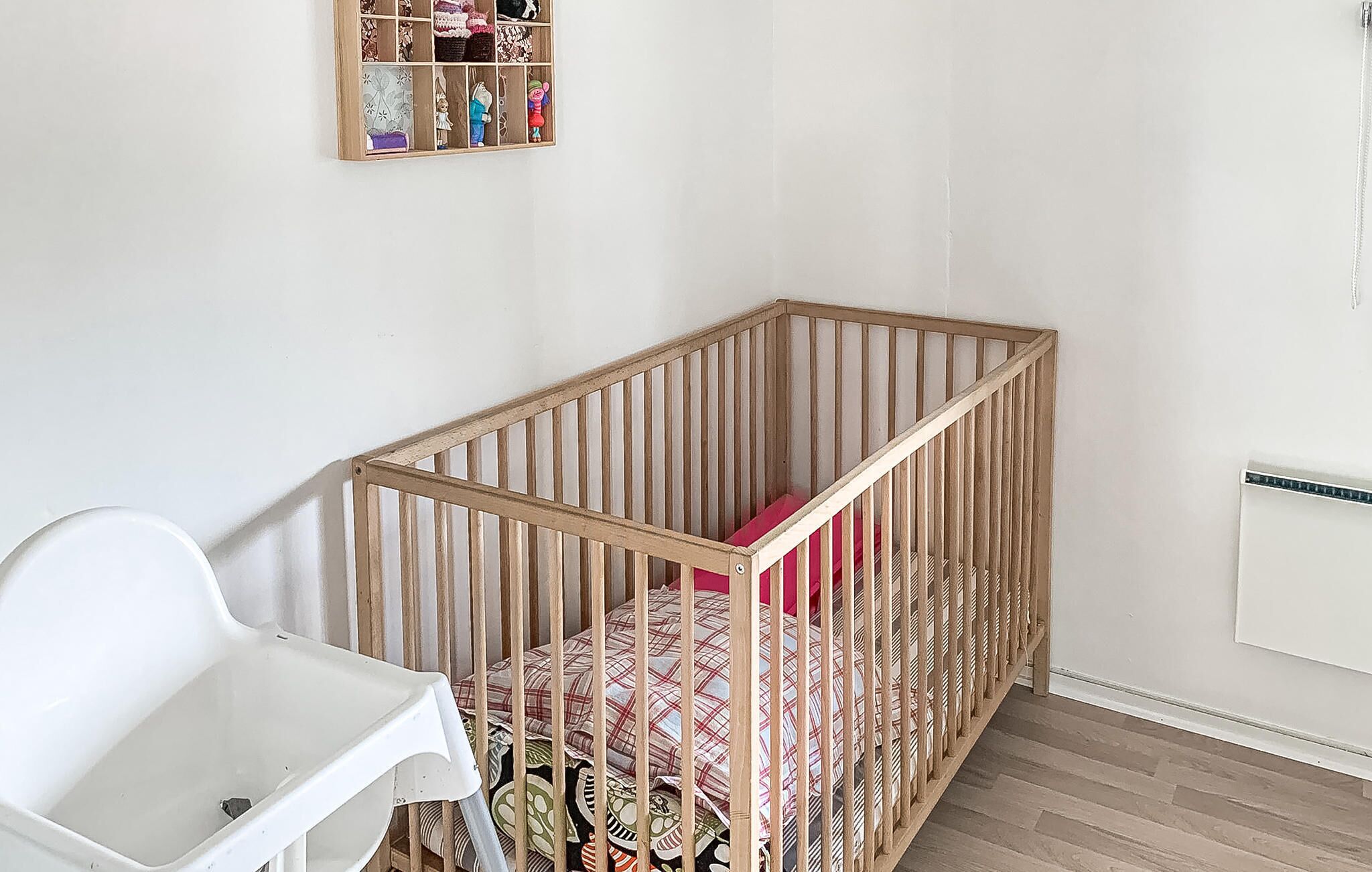 A crib with a wooden frame and a mattress with a patterned cover, a white chair, and a wall-mounted shelf with various items.