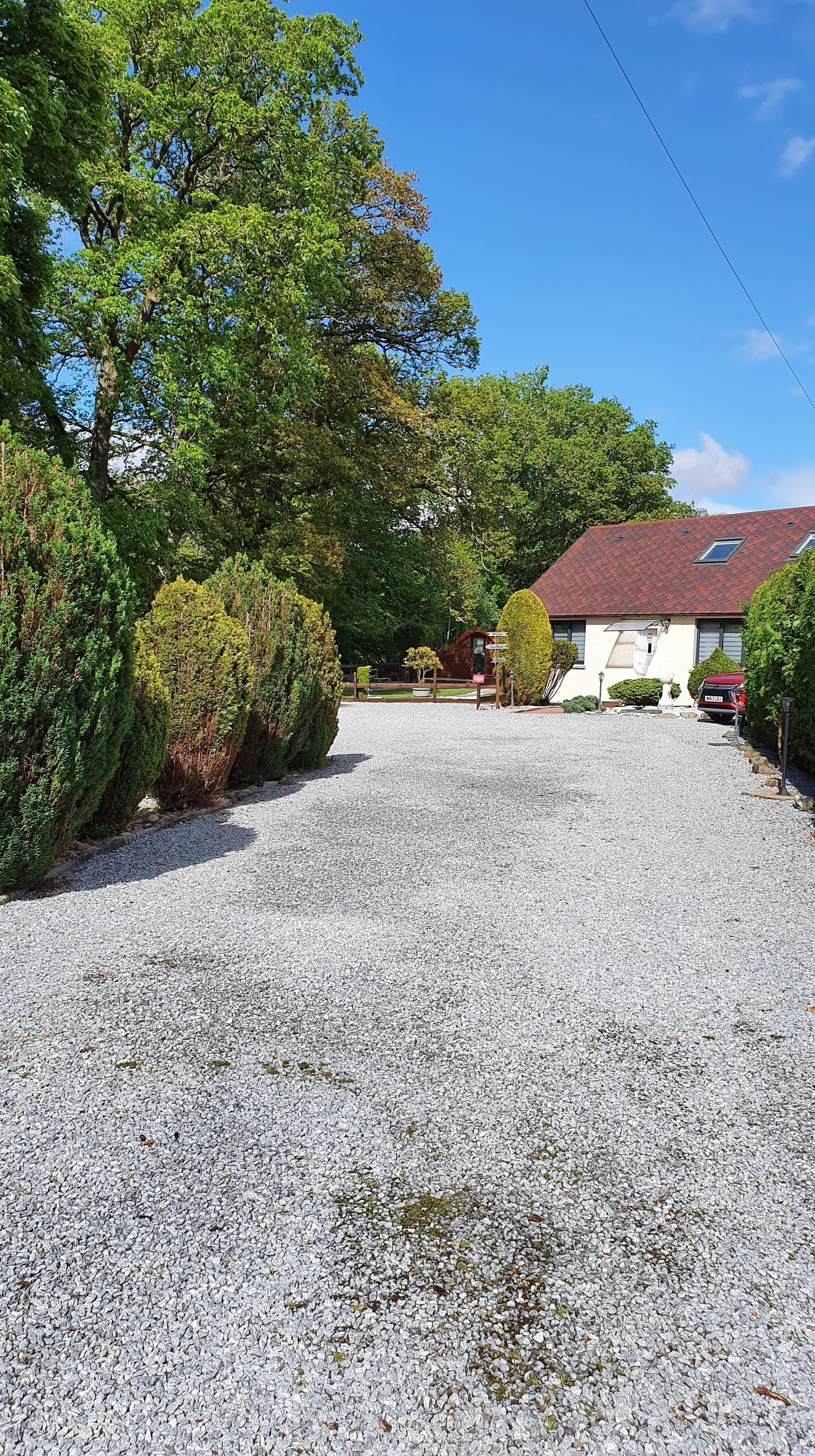 A gravel driveway leading to a house with a red roof, surrounded by well-maintained hedges and trees.
