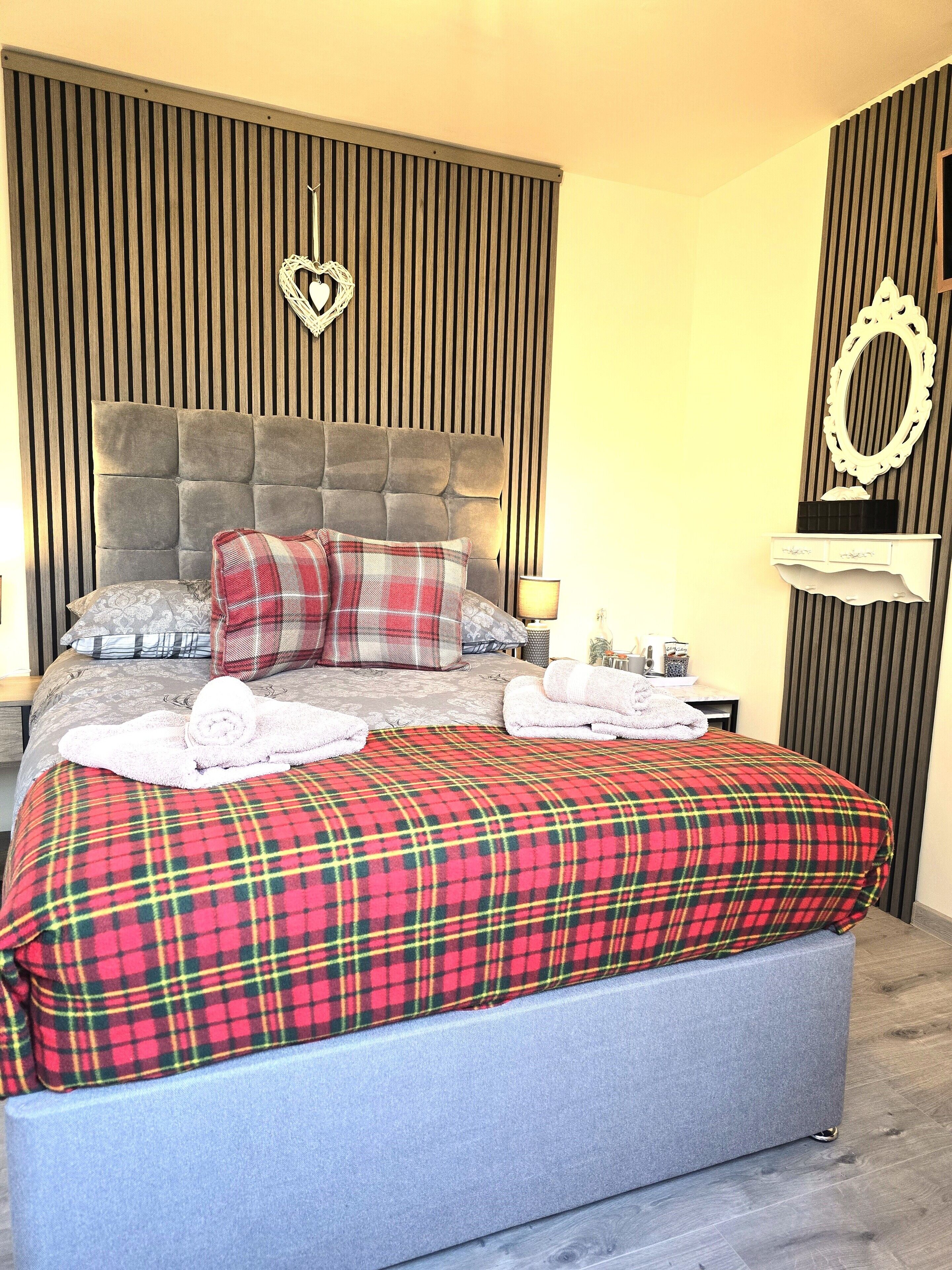 Una cama bien tendida con una colcha escocesa, un cabecero tapizado en gris y una decoración con forma de corazón encima.