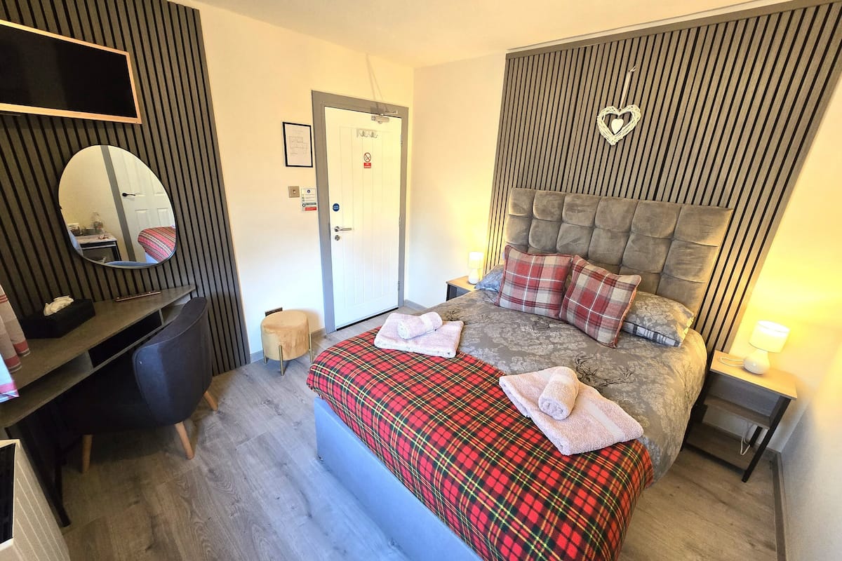 A bedroom with a bed, a bedside table, a nightstand, a mirror, a door, and a small stool.
