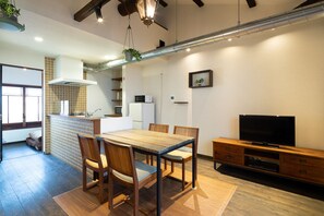A modern kitchen with a tiled backsplash, a dining area with a wooden table and chairs, a television on a wooden stand, and a hanging plant.