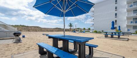 A beachside patio with a blue umbrella, picnic table, and barbecue grill.