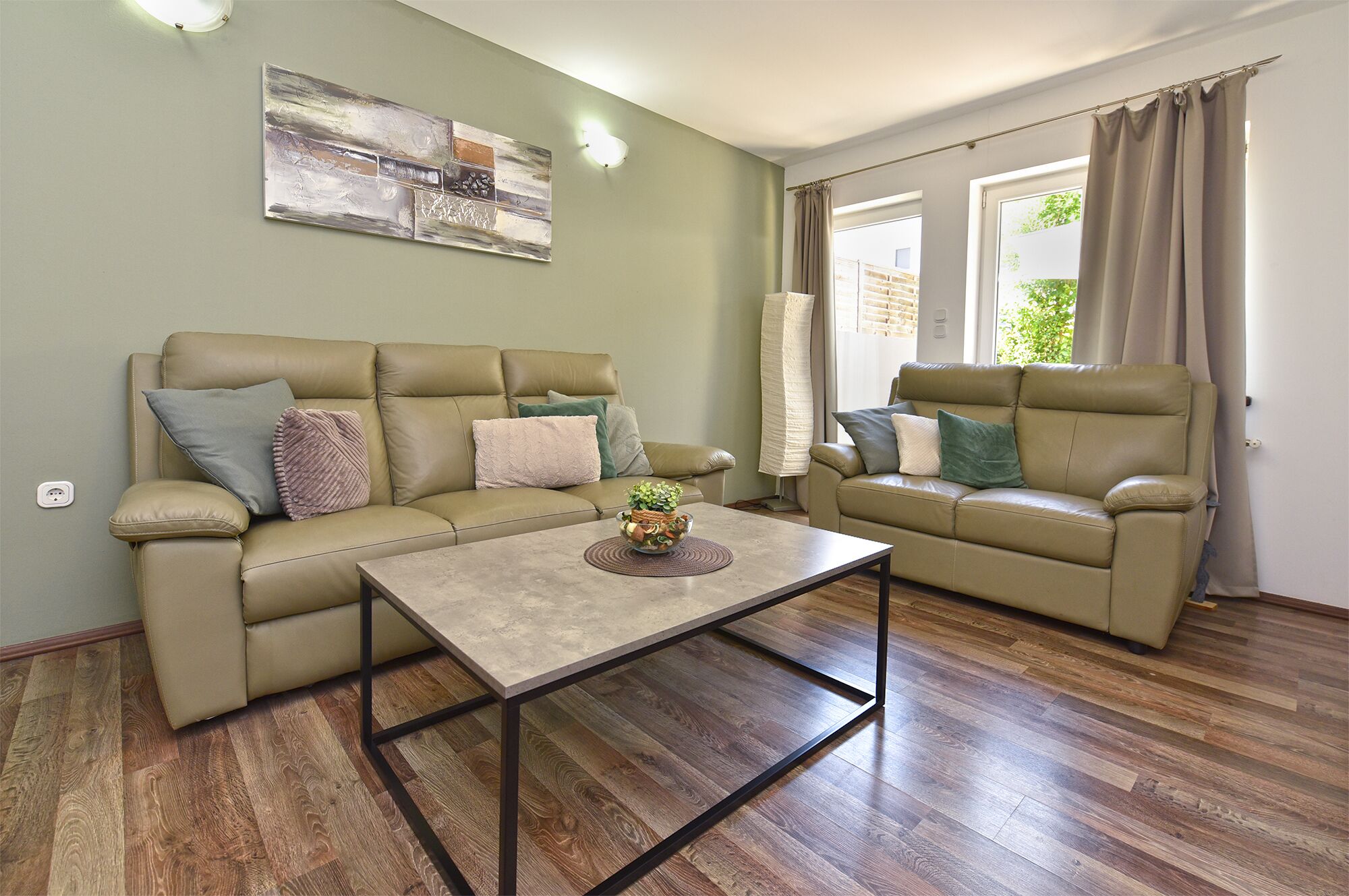 A living room with a sofa, armchair, and coffee table. There is a painting on the wall and a window with curtains.