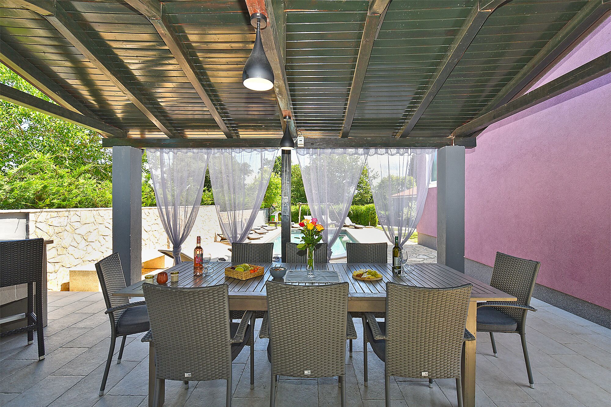 An outdoor patio with a table set for dining, surrounded by wicker chairs, under a covered area with a metal roof.
