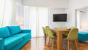 Habitación de hotel moderna con un sofá verde azulado, una mesa de comedor de madera con sillas verdes, un televisor de pantalla plana y una cama con cubierta verde azulado.