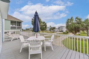 Une terrasse en bois, agrémentée de chaises Adirondack blanches et d’une table, le tout entouré d’une balustrade blanche, offre une vue sur une pelouse bien entretenue et des palmiers.