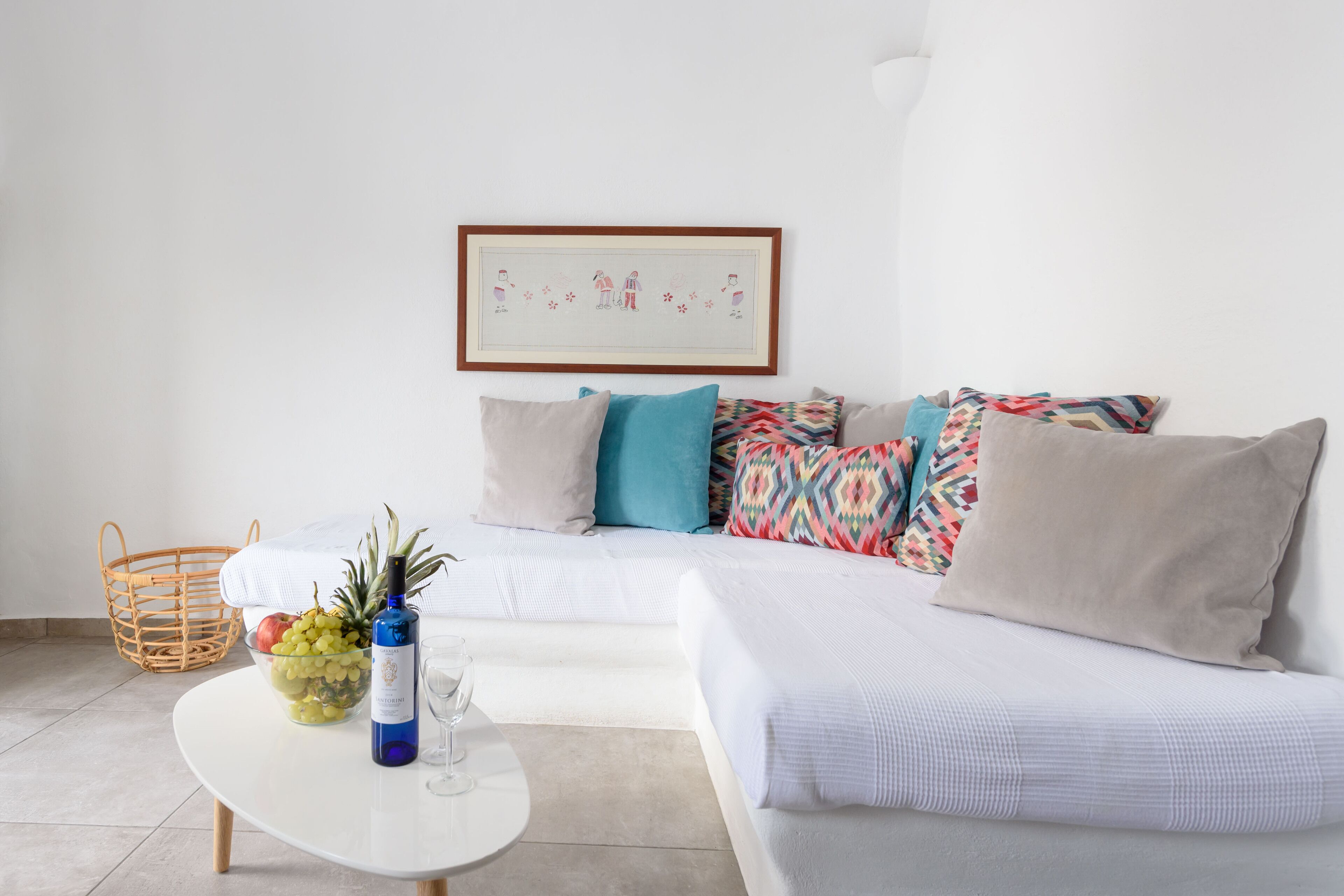 A modern living room with a white sofa, colorful pillows, a white coffee table with a fruit bowl and wine bottle, and a framed picture on the wall.