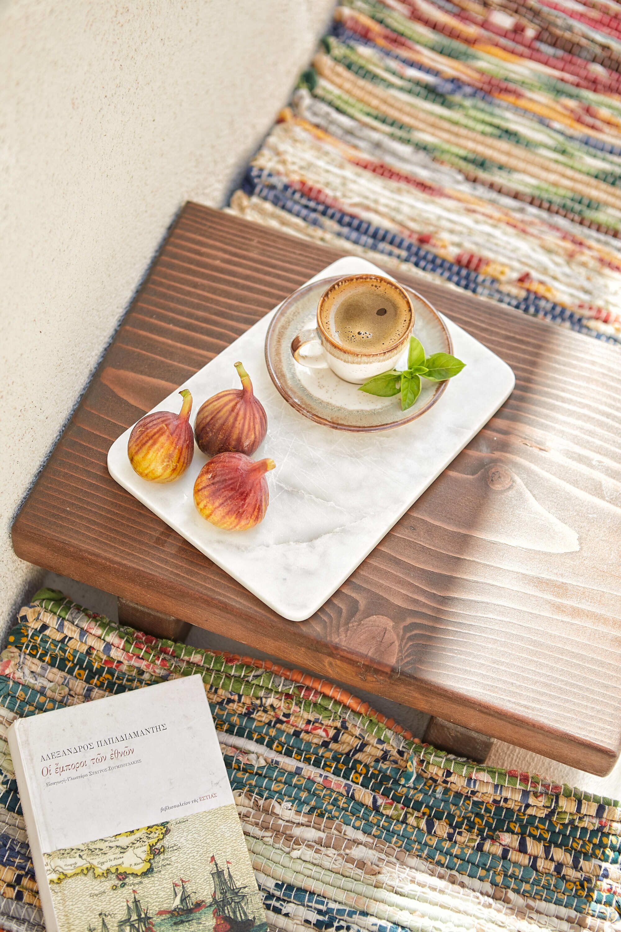 A small wooden table with a white marble coaster holding a cup and three figs. A book is placed on the table.