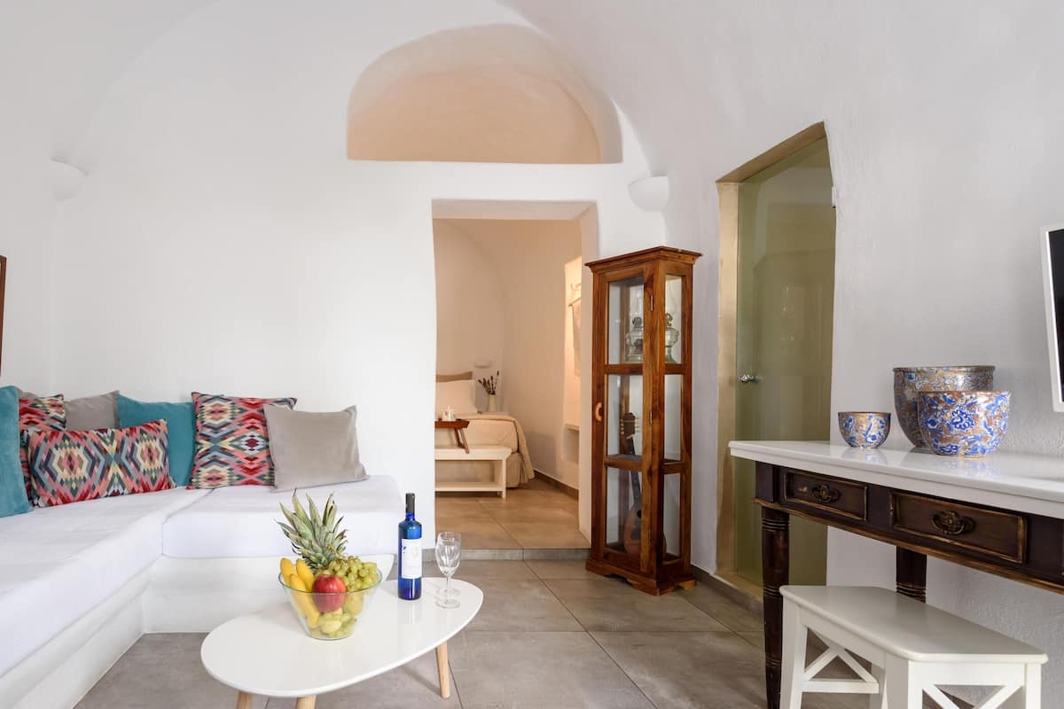 Ein modernes Wohnzimmer mit einem weißen Sofa, bunten Kissen, einer Schale mit Obst aus Glas auf einem weißen Tisch, einer Holzkommode und einem Fernseher auf einem weißen Schrank.