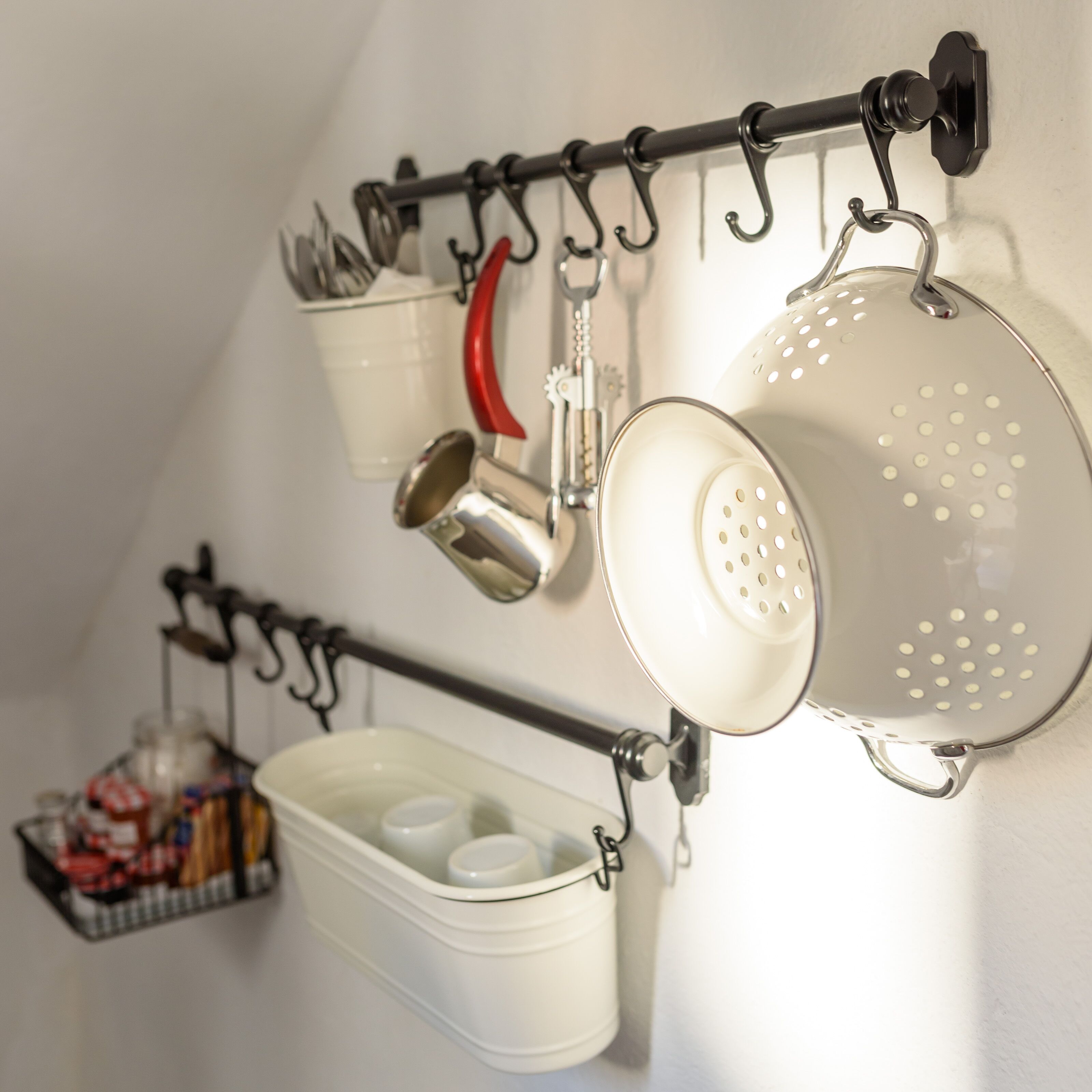 A kitchen with a utensil rack holding pots, pans, and a colander. Below, there is a white container with lids.