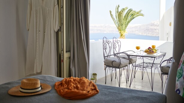 A balcony with a table set for two, a hanging dress, and a hat on a bed. - Arvanitis Cave Village (Santorini)