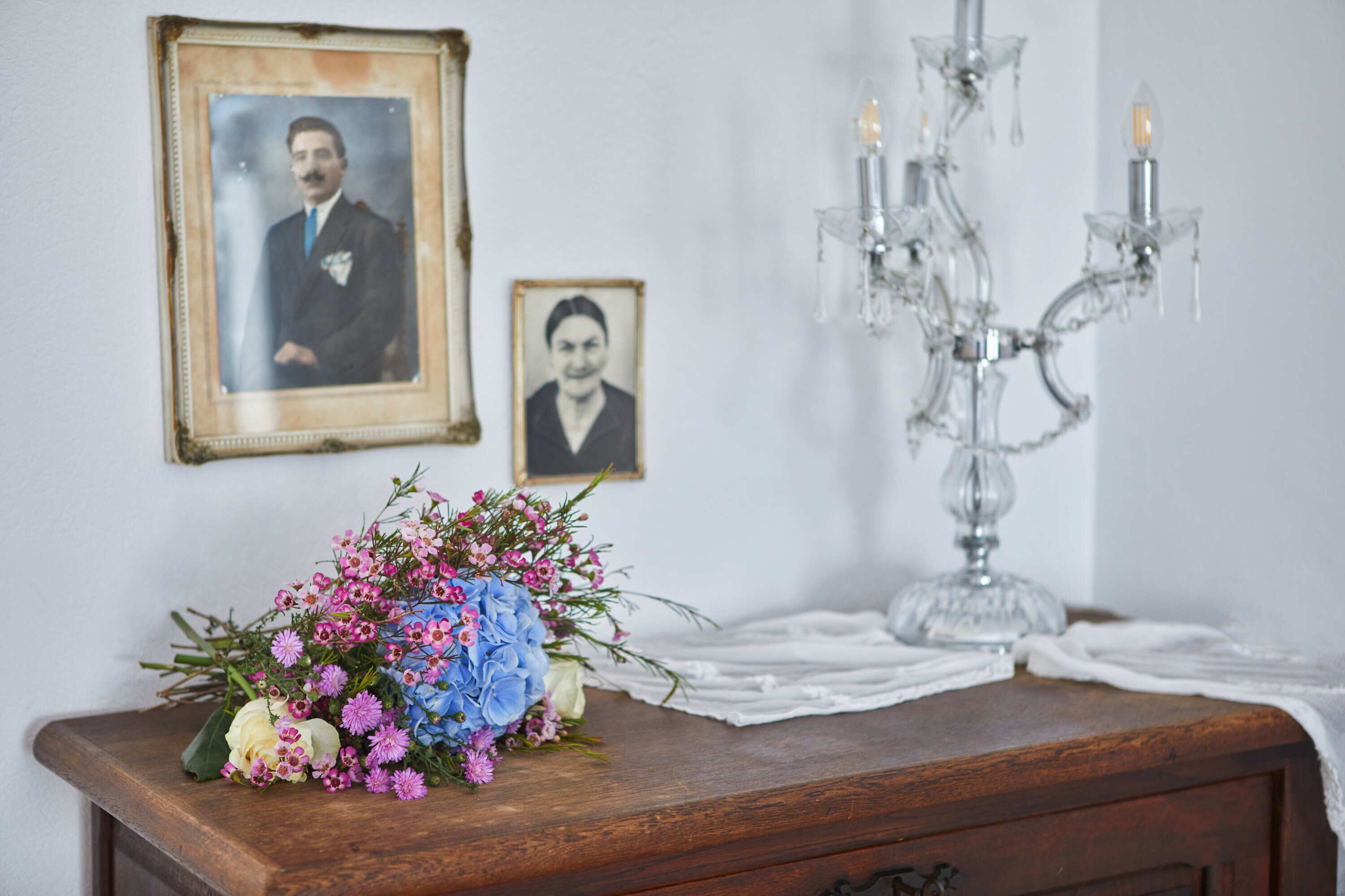 Une commode en bois ornée d’un bouquet de fleurs, de deux photographies encadrées et d’un lustre en cristal.