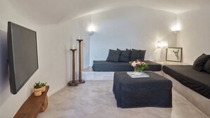 A modern, minimalist living room with a large flat-screen TV, a sofa, a coffee table with a book and flowers, and a floor lamp. - Arvanitis Cave Village (Santorini)