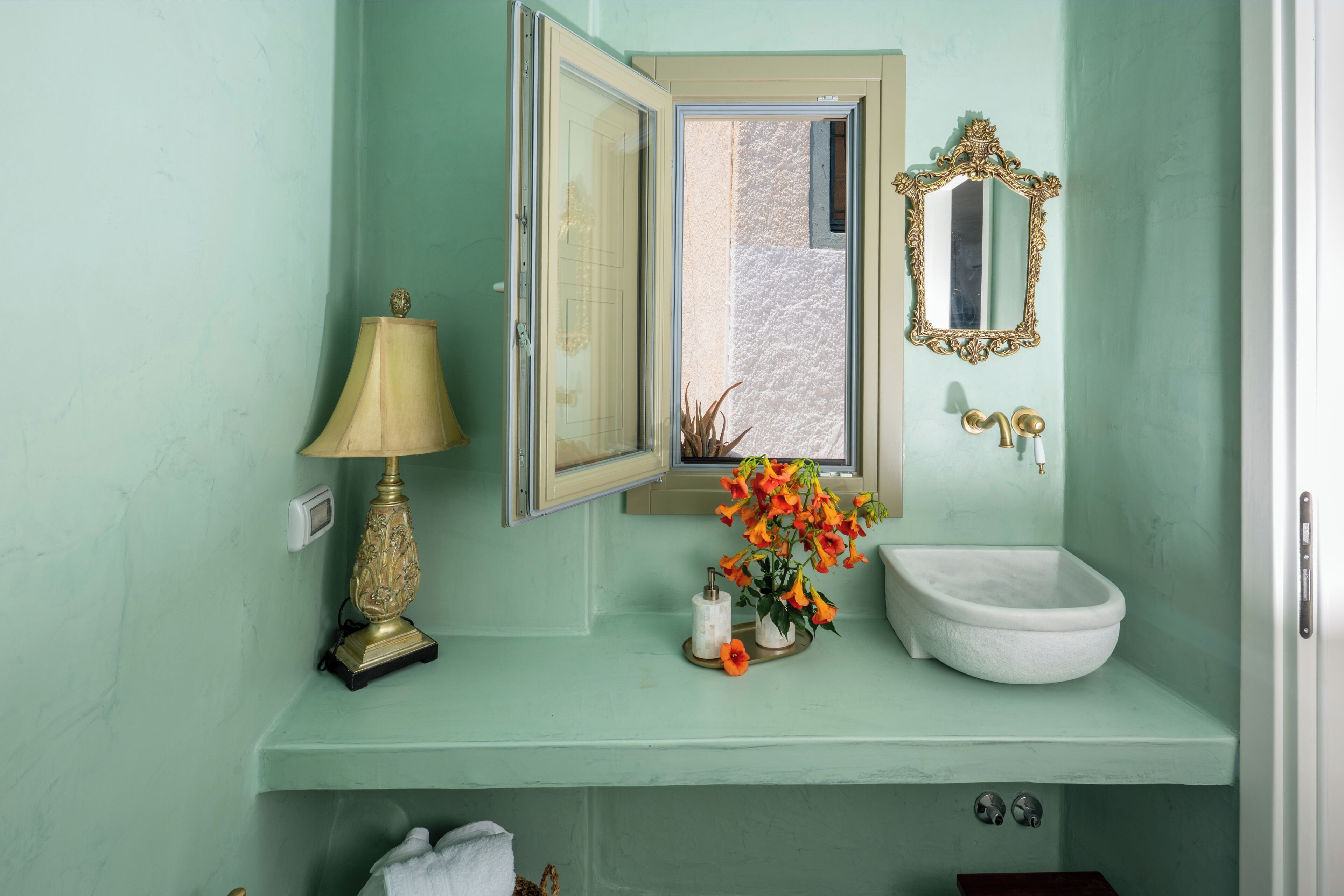Un baño con lavamanos, espejo, ventana, lámpara y un florero con flores.