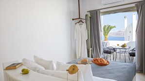 A hotel room with a bed, a small table with fruit and drinks, a balcony with a view of the sea, and a hanging garment rack. - Arvanitis Cave Village (Santorini)