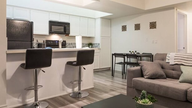 A modern kitchen with white cabinets, a black refrigerator, and a microwave. There are two black bar stools and a dining table with chairs. A gray sofa and a small potted plant are in the living area. - Cozy condo near MCC, pool, covered parking (Mesa)