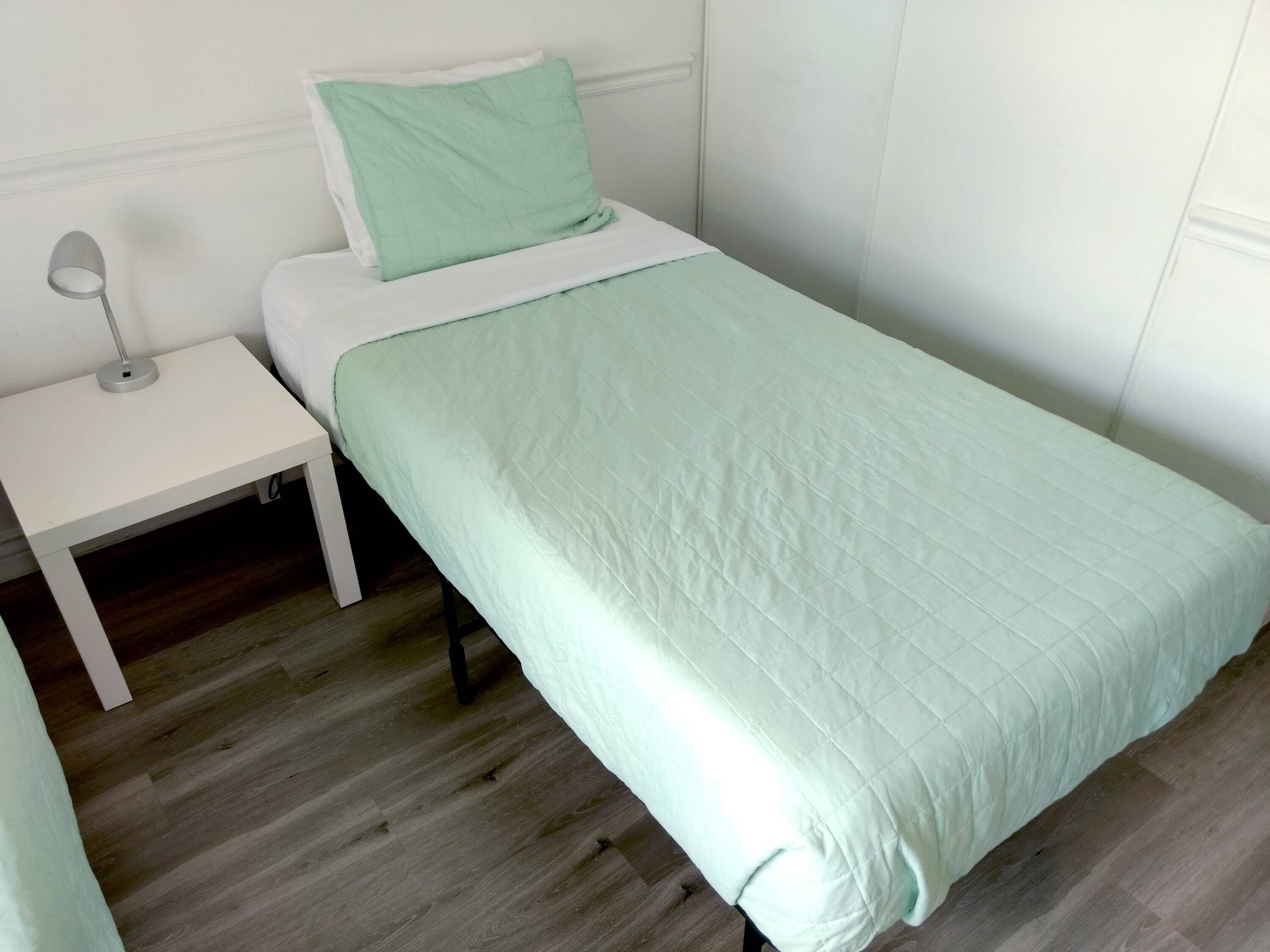 Een klein, netjes opgemaakt bed met een lichtgroene dekbedovertrek en kussen, een witte nachtkastje met een lamp, en een houten vloer.