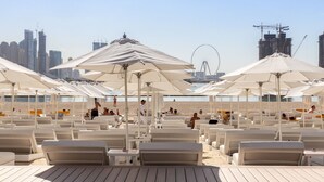 A beachside lounge area with sun umbrellas and lounge chairs, a Ferris wheel in the background, and a city skyline. - 2BR That Redefines Modern Luxury Stays at The Five Palm Jumeirah (Dubai)