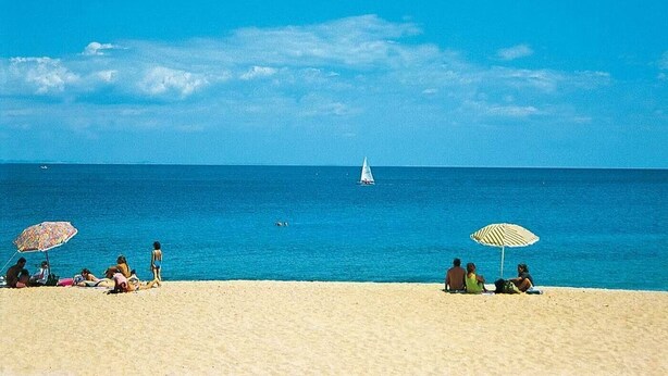 Une plage où des gens se prélassent au soleil, avec deux parasols et un voilier à l’horizon.